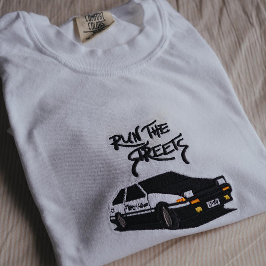 White t-shirt with black graphic design of a car and text 'Run the Streets' on a beige surface.